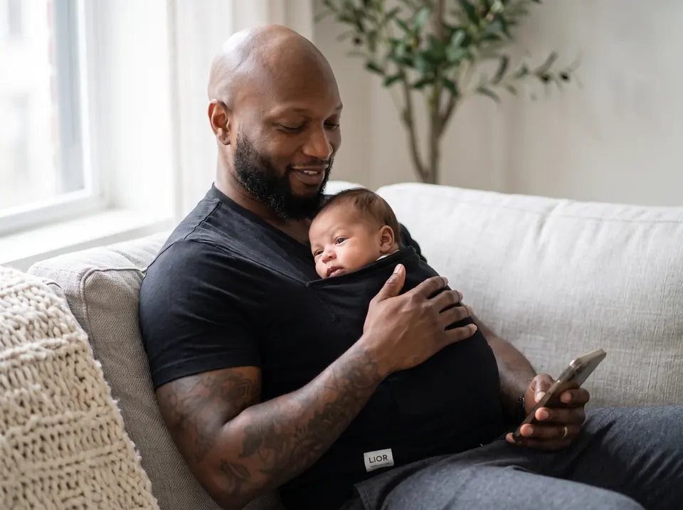 Father relaxing on a sofa with his newborn awake in the pocket of his black LIOR shirt, one hand on the baby and his phone in the other